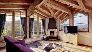 Gemütliches Wohnzimmer im Dachgeschoss mit Holzbalken, Kamin und Bergblick durch große Fenster.