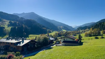 Malerische Alpenlandschaft mit grünen Hügeln, traditionellen Häusern und schneebedeckten Gipfeln in der Ferne.