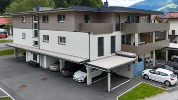 Modernes Mehrfamilienhaus mit Holzfassade, Balkonen und überdachten Parkplätzen in alpiner Umgebung.