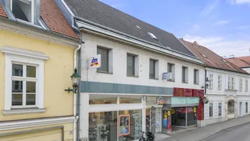 Fassade eines Geschäftsgebäudes mit Schaufenstern und angrenzenden historischen Gebäuden an einer Straße.