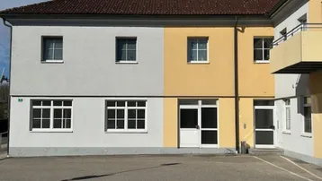 Fassade eines Gebäudes mit zwei Farbbereichen, hellgrau und gelb, unter einem traditionellen Ziegeldach.