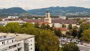 Weitläufiger Blick über die Stadt mit grünen Hügeln und einem Kirchturm im Hintergrund.