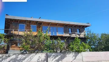 Holzverkleidetes Gebäude mit großen Fenstern und einem Betonzaun im Vordergrund unter blauem Himmel.