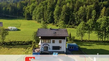 Freistehendes Haus in ländlicher Umgebung mit grünen Wiesen und bewaldeten Bergen im Hintergrund.