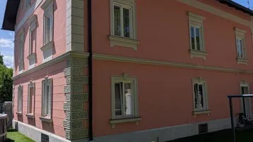 Historisches Gebäude mit rosafarbener Fassade, weißen Fensterrahmen und gepflegtem Rasen unter blauem Himmel.