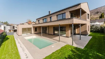 Modernes Holzhaus mit großem Pool, Rasenfläche und Bergblick unter blauem Himmel.