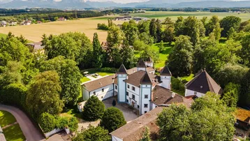 Luftaufnahme eines historischen Anwesens mit Türmen, umgeben von viel Grün und mit Bergblick.