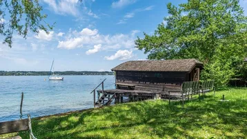 Altes Holzbootshaus am Seeufer mit Steg und Blick auf den See unter blauem Himmel.