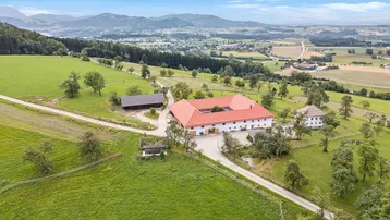 Luftaufnahme eines weitläufigen Anwesens mit mehreren Gebäuden, umgeben von grünen Feldern und Bergen.