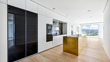 Moderne Küche mit weißer Front, goldener Kücheninsel und Holzboden, bietet weite Aussicht.