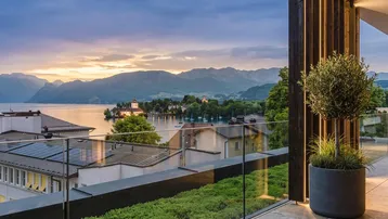 Geräumiger Balkon mit Glasgeländer und atemberaubendem Blick auf den See und die umliegenden Berge bei Sonnenuntergang.