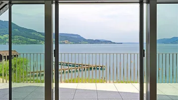 Blick auf den See von einem Innenraum mit großer Glasschiebetür und Terrasse.