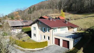 Einfamilienhaus in ländlicher Umgebung mit Doppelgarage und Bergblick unter blauem Himmel.