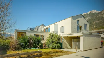 Modernes Gebäude mit heller Fassade, Gartenbereich und Bergblick unter blauem Himmel.