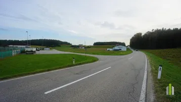 Landstraße führt durch grüne Felder und Wälder, mit Gebäuden in der Ferne unter bewölktem Himmel.