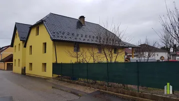 Gelbes Wohnhaus mit dunklem Ziegeldach und grüner Umzäunung an einem bewölkten Tag.