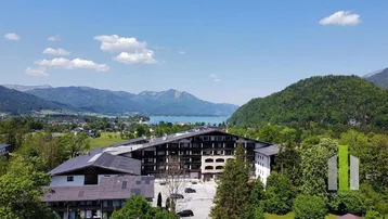Luftaufnahme eines Gebäudekomplexes mit See- und Bergblick in grüner Umgebung unter blauem Himmel.