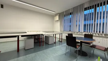 Heller Büroraum mit großen Fenstern, Jalousien und funktionalen Tischen für Besprechungen oder Arbeitsplätze.