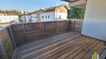 Geräumiger Balkon mit Holzboden und Holzgeländer, bietet einen Blick auf die umliegenden Gebäude und Grünflächen.
