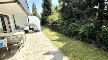 Geräumige geflieste Terrasse mit Gartenmöbeln und angrenzendem Rasen, umgeben von üppigem Grün.