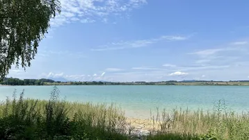 Idyllischer Blick auf einen ruhigen See mit grünen Ufern und blauem Himmel an einem sonnigen Tag.