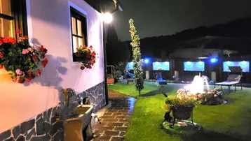 Nächtlicher Gartenbereich mit beleuchtetem Brunnen, Liegestühlen und blühenden Blumenkästen am Haus.