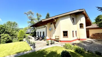Charmantes Haus mit großer Holzterrasse, umgeben von einem grünen Garten und Holzstapel.