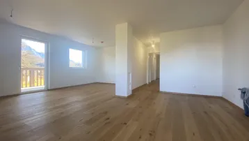 Heller, leerer Wohnbereich mit Holzboden, großen Fenstern und Zugang zum Balkon mit Bergblick.