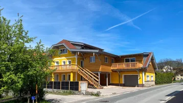 Neues Gebäude mit gelber Fassade, Holzverkleidung, Balkonen und Photovoltaikanlage unter blauem Himmel.