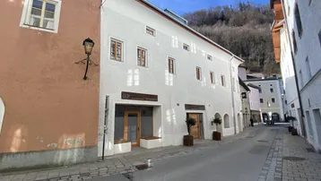 Historisches Gebäude in einer Gasse mit weißer Fassade und traditionellen Fenstern, umgeben von Bergen.
