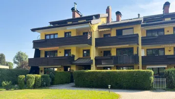 Gelbes Mehrfamilienhaus mit mehreren Balkonen und gepflegtem Vorgarten unter blauem Himmel.
