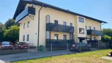 Mehrfamilienhaus mit mehreren Balkonen und Parkplätzen im Freien unter blauem Himmel.