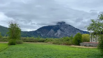 Grüne Wiesen und bewaldete Hügel mit einem majestätischen Berg im Hintergrund unter bewölktem Himmel.