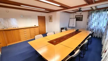 Geräumiger Konferenzraum mit großem Holztisch, blauer Teppichboden und sichtbaren Deckenbalken, ideal für Besprechungen.