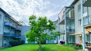 Mehrfamilienhaus mit begrüntem Innenhof, Bäumen und Balkonen unter bewölktem Himmel.
