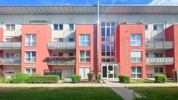 Modernes Mehrfamilienhaus mit roter Fassade, Balkonen und großen Fenstern, umgeben von Grünflächen.