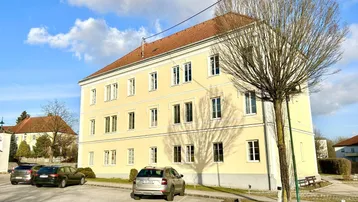 Gelbes Mehrfamilienhaus mit rotem Dach und Parkplätzen unter blauem Himmel.