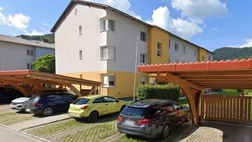 Mehrfamilienhaus mit gelb-weißer Fassade und überdachten Parkplätzen im Vordergrund unter blauem Himmel.