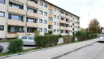 Mehrfamilienhaus mit Balkonen und Parkplätzen im Freien, umgeben von gepflegten Büschen.