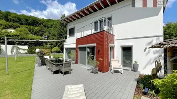 Große Holzterrasse mit Esstisch und Liegestühlen vor dem modernen Haus mit roter Akzentfassade.