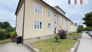 Mehrfamilienhaus mit beiger Fassade, weißen Fenstern und kleinem Vorgarten an einer Straße.