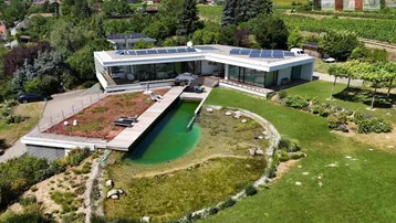 Moderne Villa mit großem Naturpool, Holzdeck und Photovoltaikanlage auf dem Dach in grüner Landschaft.