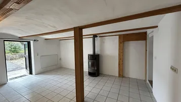 Geräumiger Innenraum mit Fliesenboden, Holzbalken und einem modernen Holzofen.