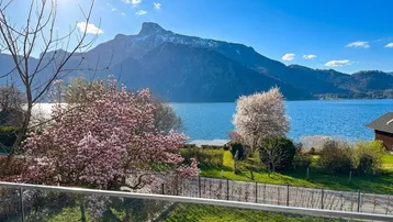 Blick vom Balkon auf blühende Bäume, den See und die umliegende Berglandschaft.