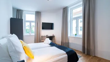 Helles Schlafzimmer mit Doppelbett, Holzboden und zwei großen Fenstern, die viel Tageslicht hereinlassen.