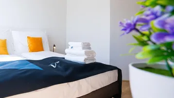 Modernes Schlafzimmer mit Doppelbett, frischen Handtüchern und einem Farbtupfer durch orange Kissen.