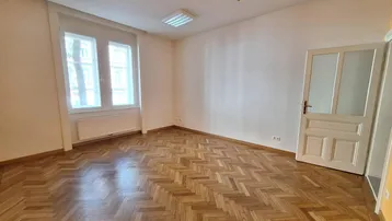 Geräumiger, leerer Raum mit Fischgrätparkettboden, großen Fenstern und einer weißen Tür.