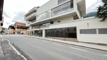 Modernes Gebäude mit großen Glasfassaden und Balkonen an einer Straße, umgeben von Bergen.