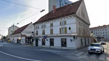 Historisches Gebäude an einer Straßenecke mit traditionellen Fensterläden und Ziegeldach in städtischer Umgebung.