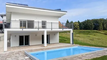 Modernes weißes Haus mit Swimmingpool und großer gepflasterter Terrasse, umgeben von grünen Hügeln.